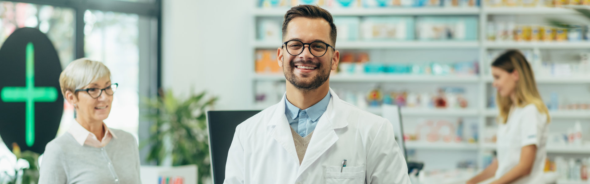 Young female pharmacist posing while working in a pharmacy while his female colleague is selling prescription medications to senior female customer